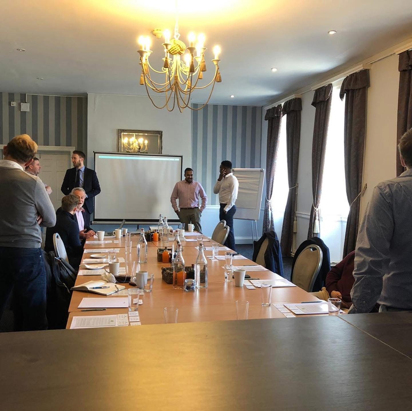 A long meeting table set for a gathering of business people under a chandelier, with guests talking in small groups